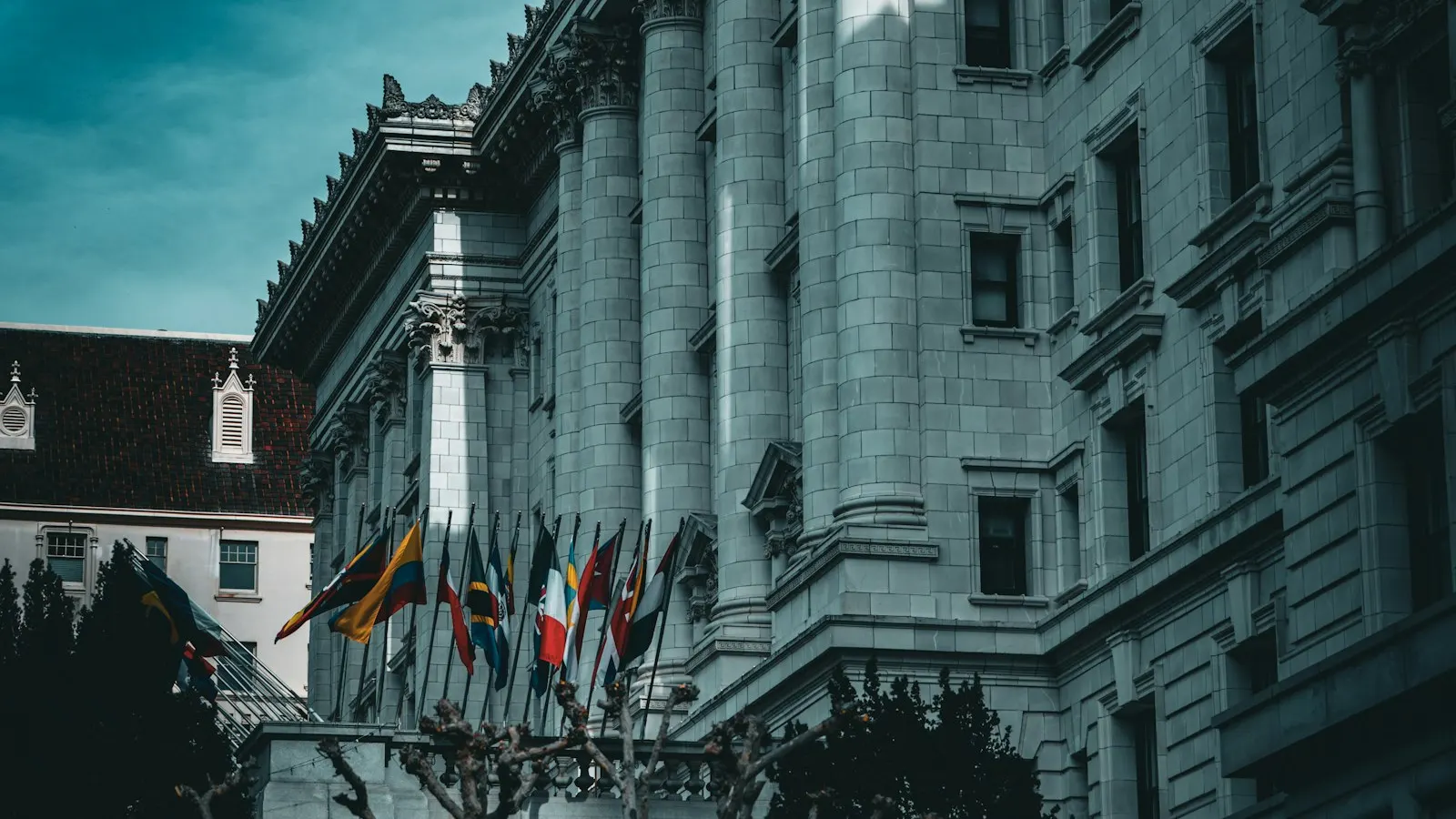 A row of national flags on angled poles along the stone facade of a columned government building under a dark teal sky