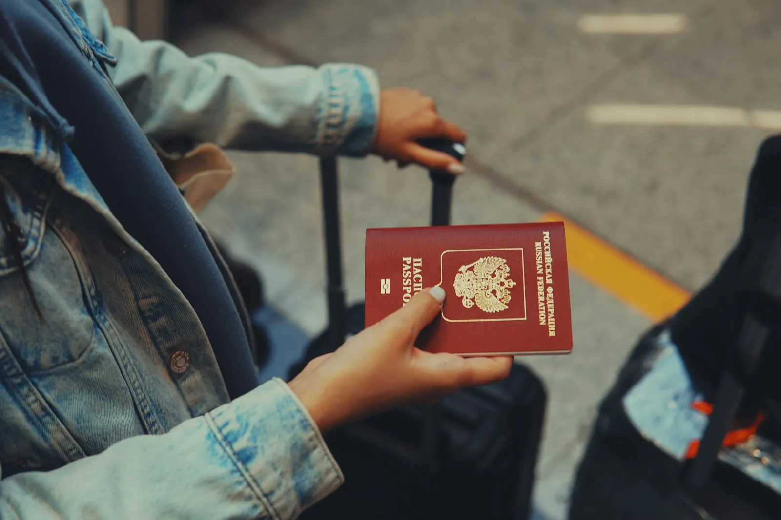 A traveler in a denim jacket holding a red Russian Federation passport next to a suitcase