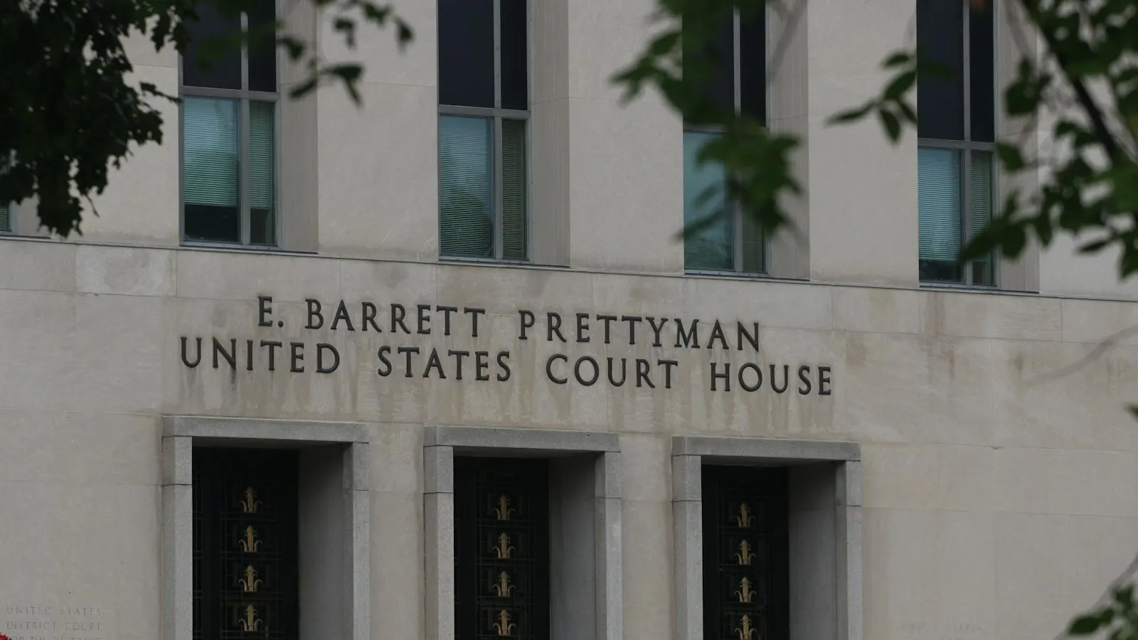 The limestone facade of the E. Barrett Prettyman United States Court House in Washington, DC, its name carved above a row of tall narrow windows and framed by overhanging tree branches