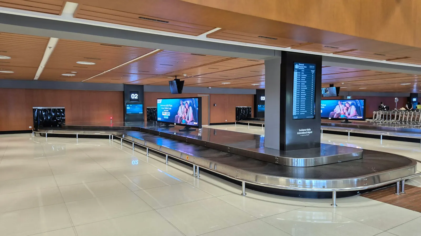 An empty airport baggage claim hall with two curved carousels under a timber-panelled ceiling, information screens mounted on a pillar between them and rows of seating along the far wall