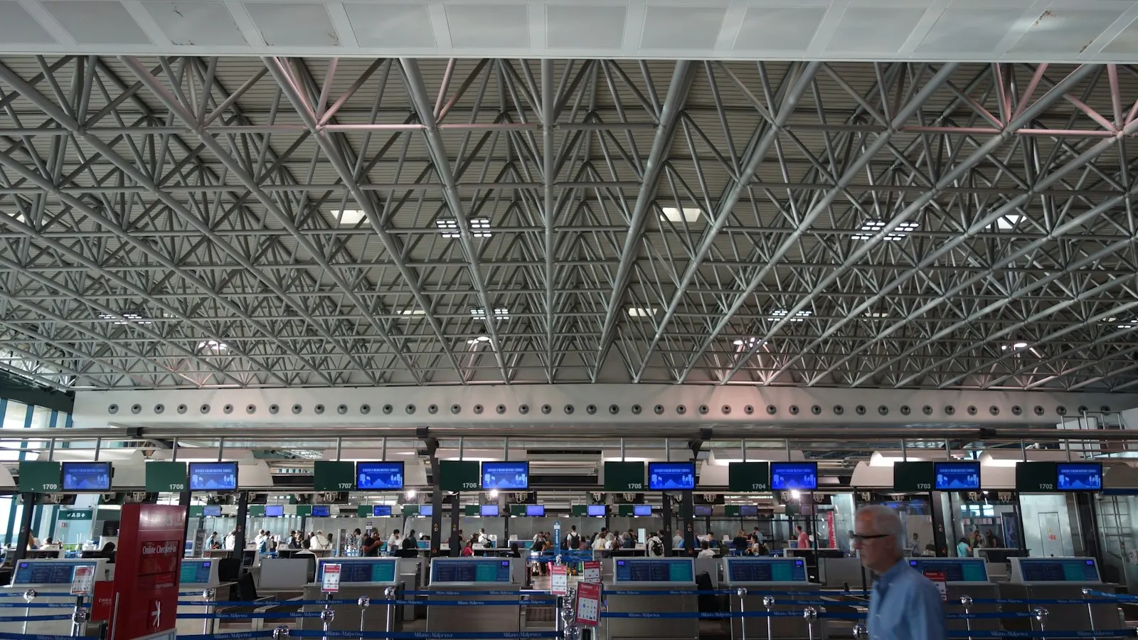 A long row of airline check-in desks under a steel truss roof, passengers queuing at the counters with overhead screens and numbered bay signs above each desk
