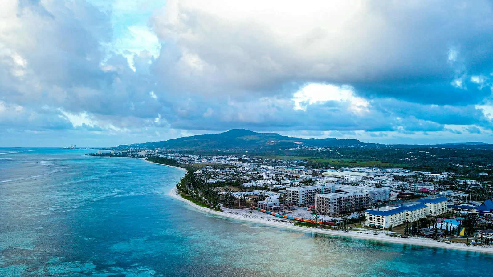 An aerial view of a tropical coastal town, hotel and apartment blocks lining a pale beach with a turquoise reef flat in the foreground and a green ridge rising behind under heavy cloud