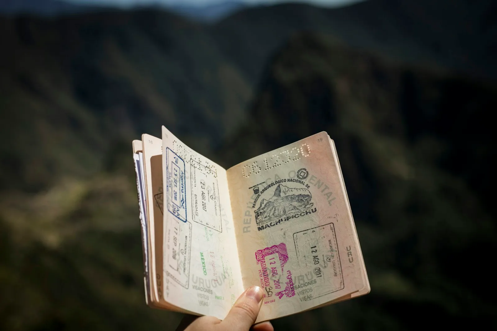 A hand holding up an open passport full of entry stamps, including a Machu Picchu stamp, against a blurred mountain backdrop