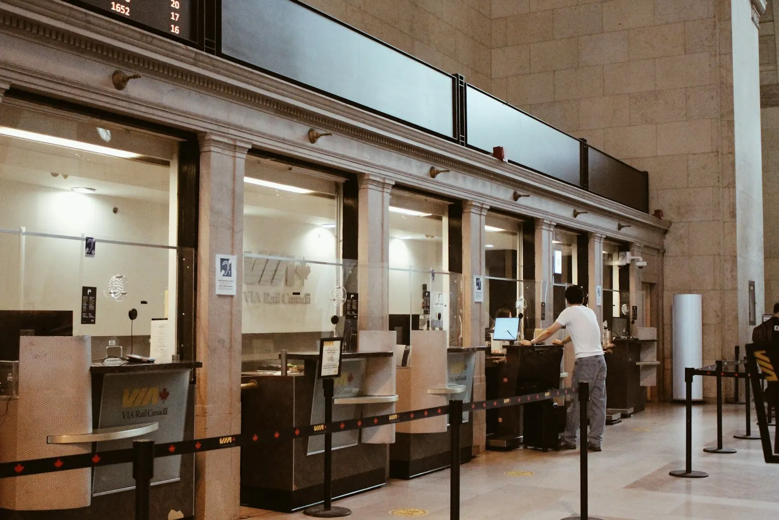 A traveler standing at a ticket counter in a large stone hall, with a line of service windows and queue barriers beside him